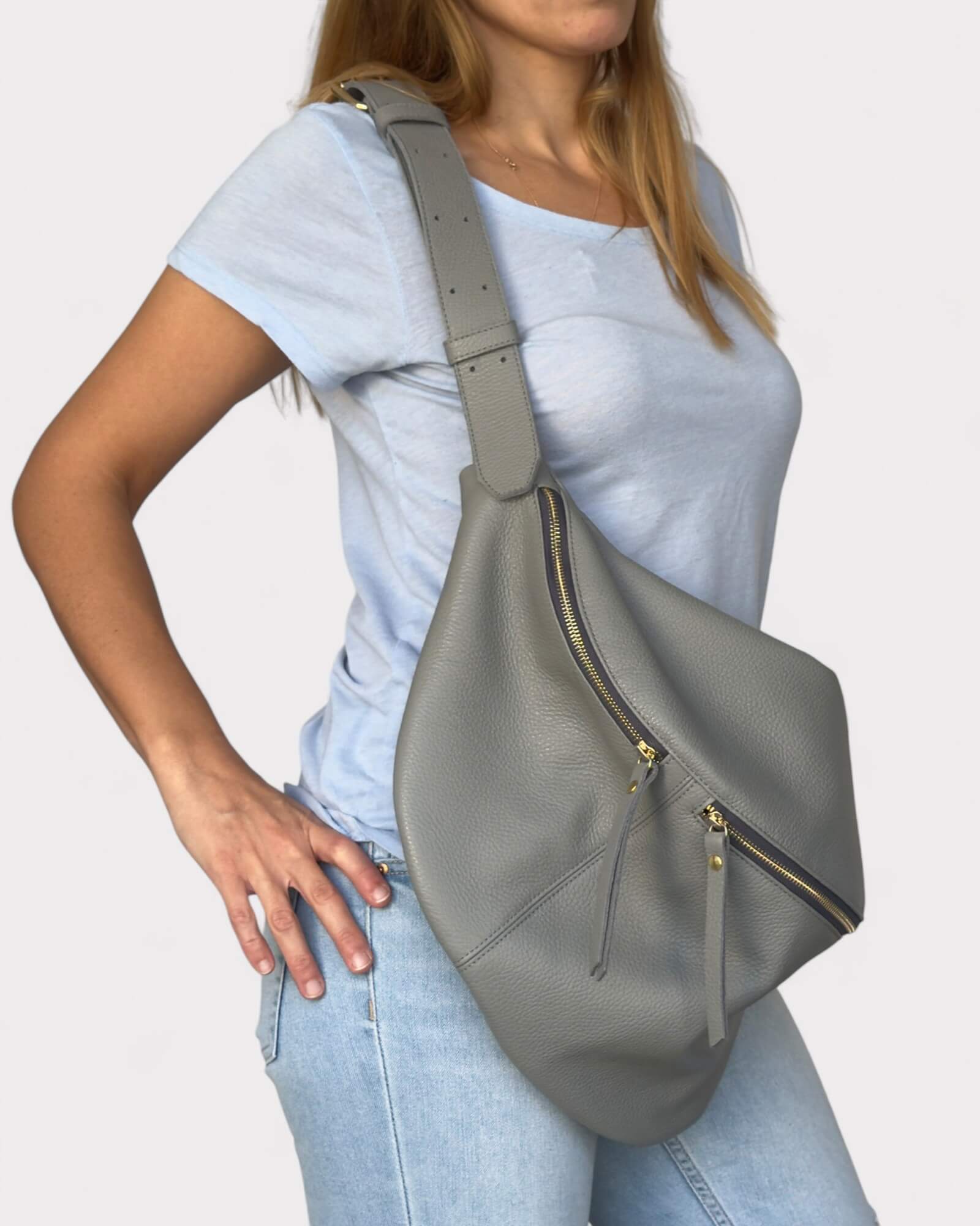 Person wearing a gray sling bag with gold zippers on a plain background