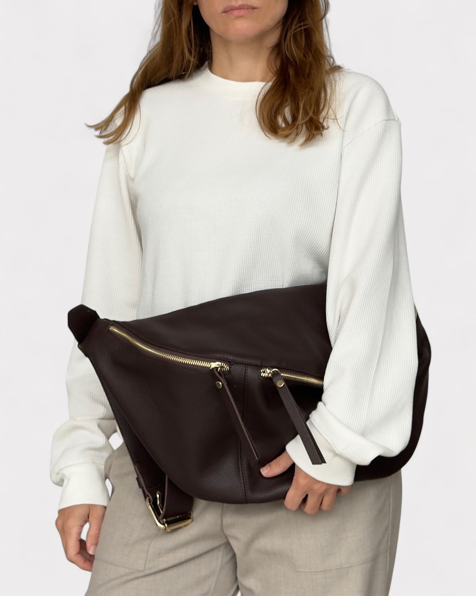 Person wearing a white sweatshirt and beige pants with a brown leather bag over their shoulder on a plain background