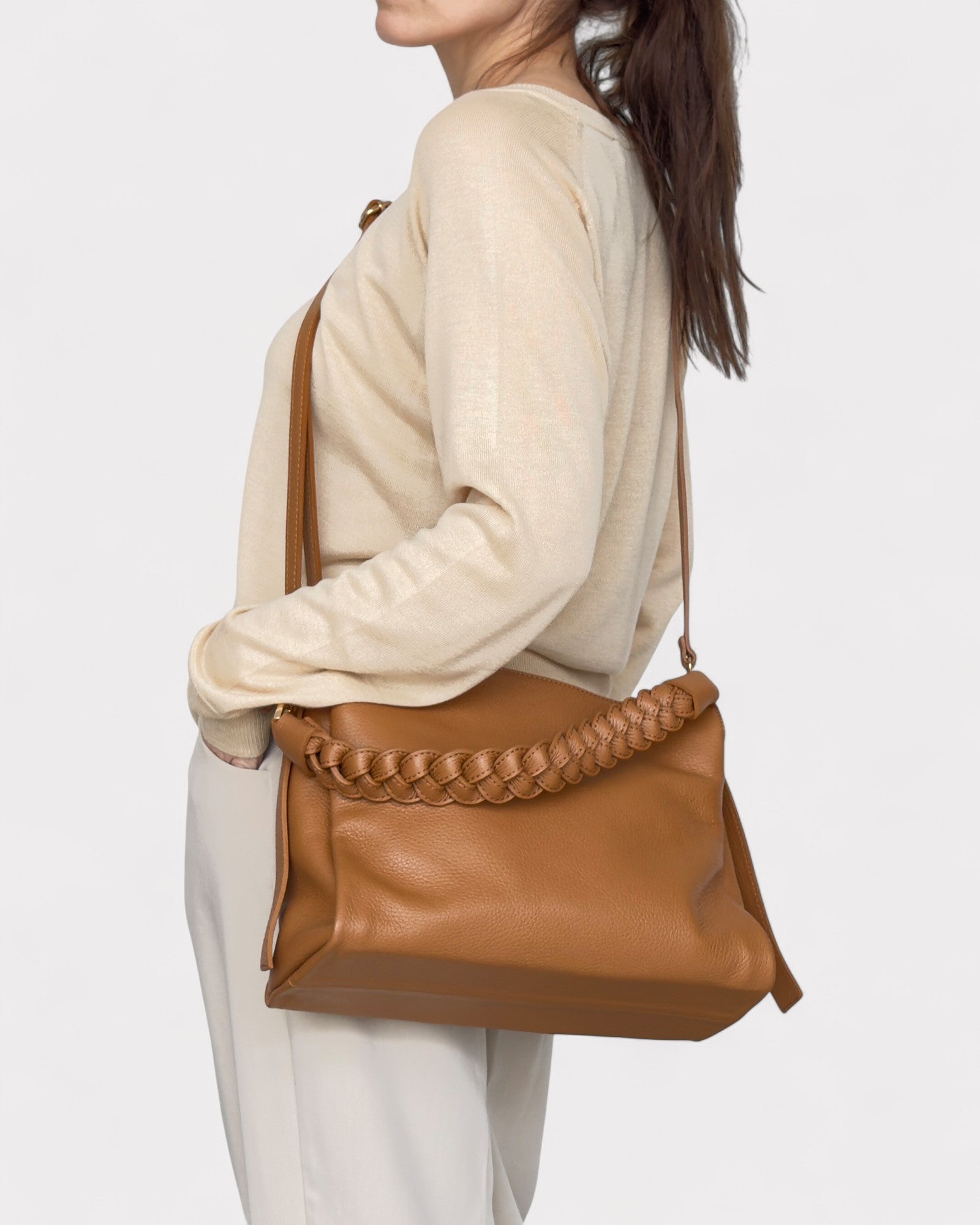 Person wearing a beige sweater and white pants with a Tan leather handbag featuring braided details on a light gray background.