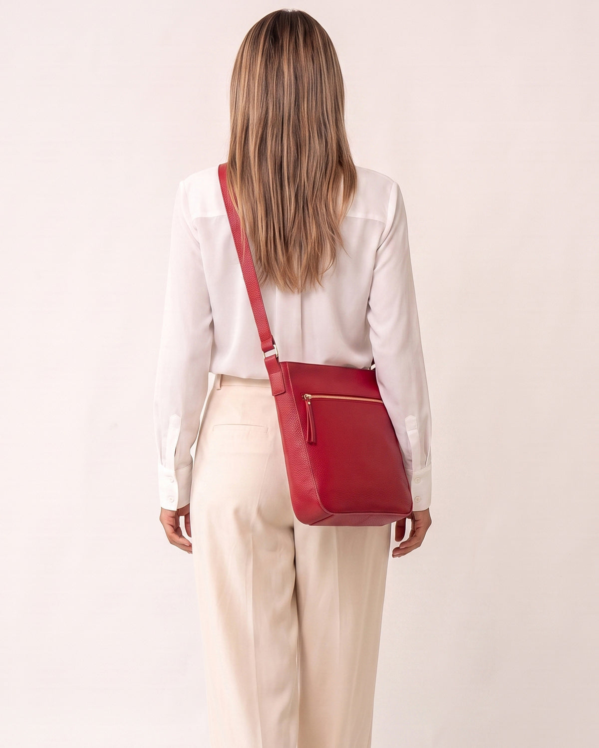 A woman viewed from behind wearing a white blouse and cream trousers, carrying a red leather crossbody bag with a gold zipper pull and gold hardware against a plain off-white background.