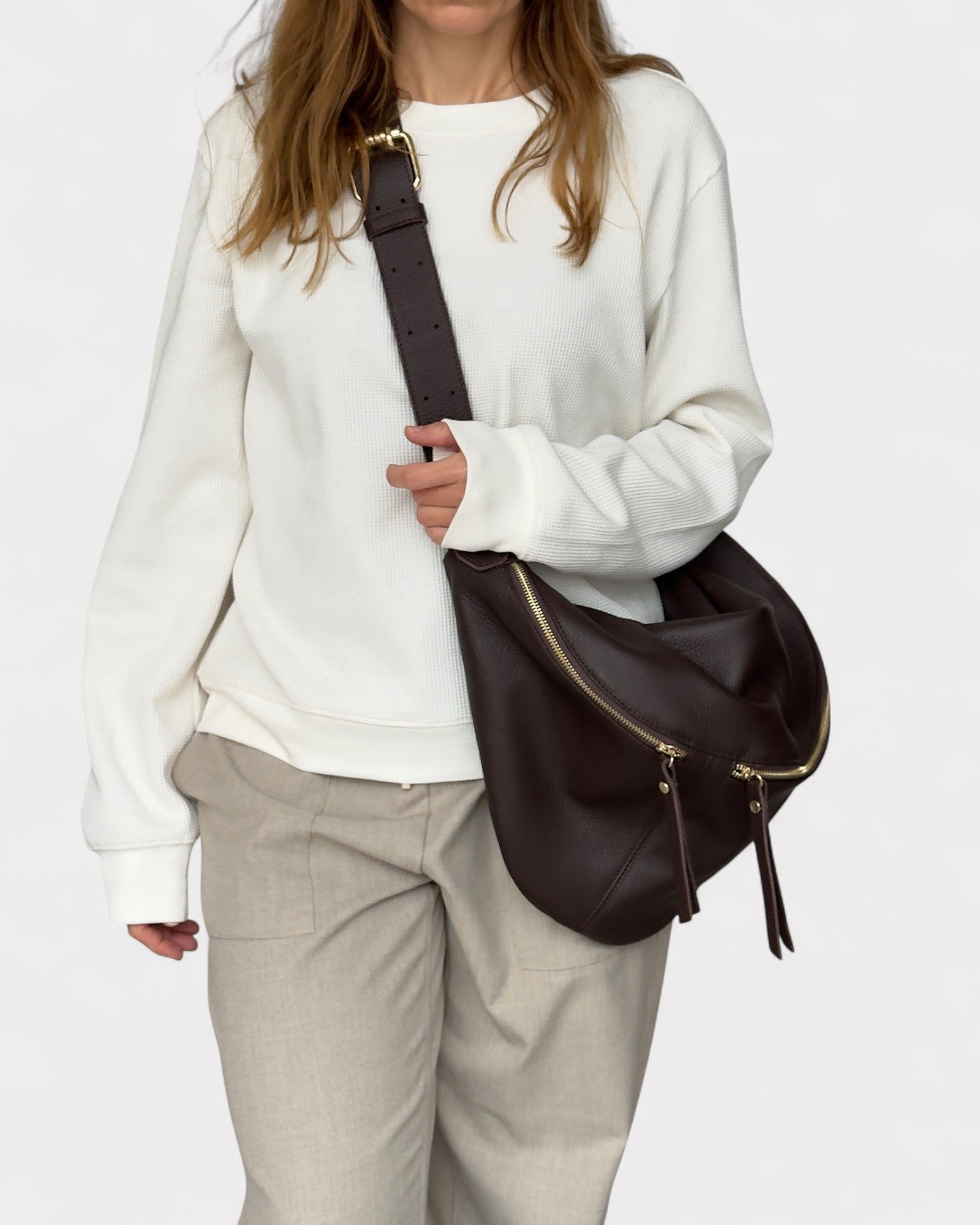 Person wearing a white sweater and beige pants holding a brown leather bag on a white background