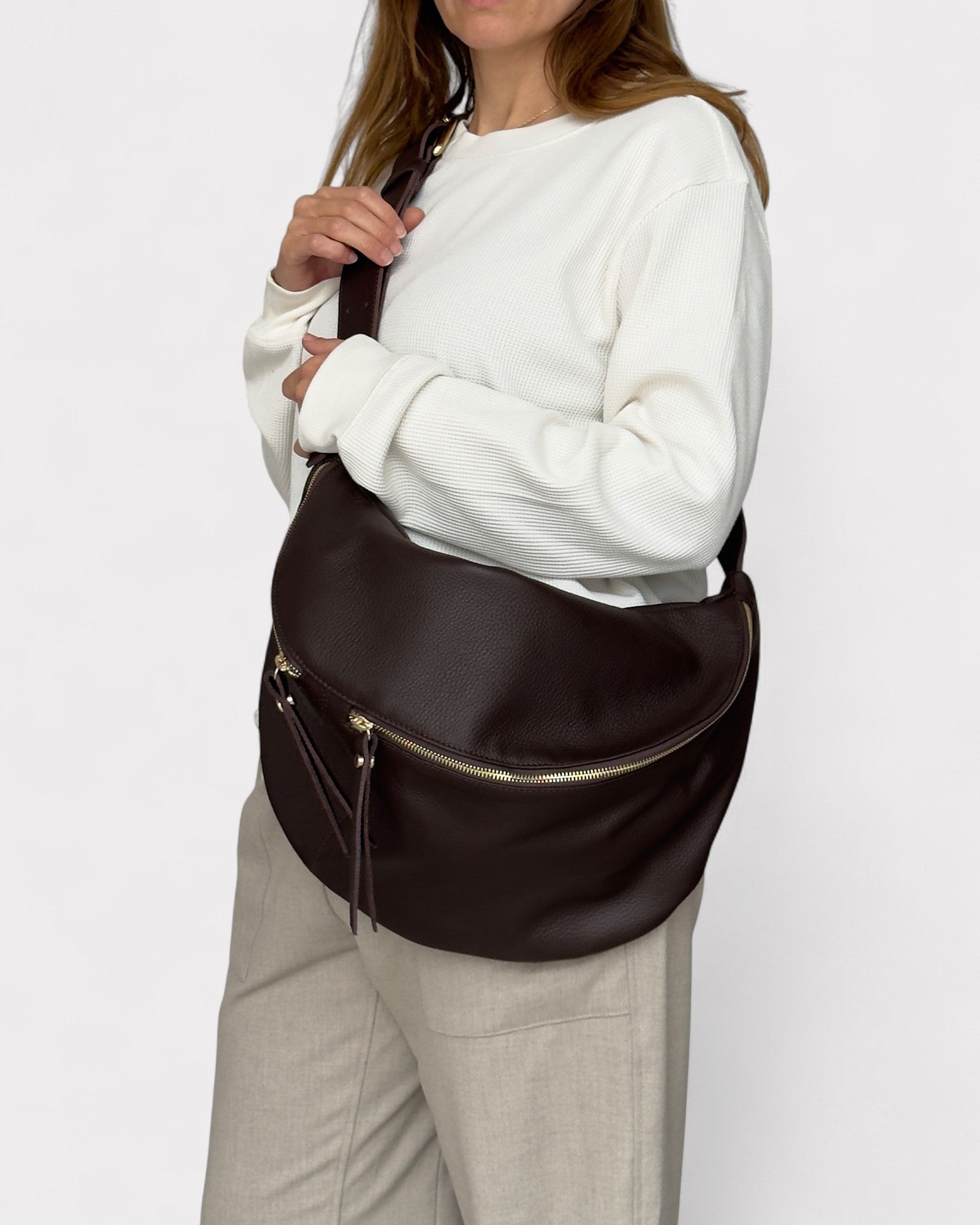 Person wearing a white sweater and beige pants holding a brown leather bag on a white background